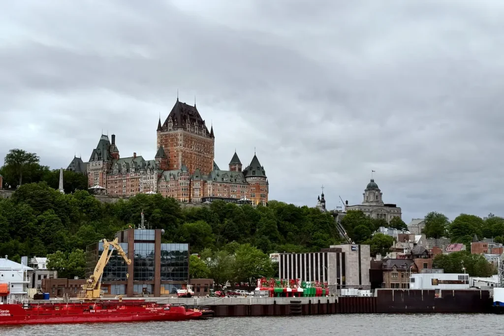 The Château Frontenac towers over Old Québec City with its distinctive green copper rooftops and central spire, set against a cloudy gray sky. Below, a modern waterfront area features a bright red Canadian Coast Guard ship and industrial buildings along the harbor.