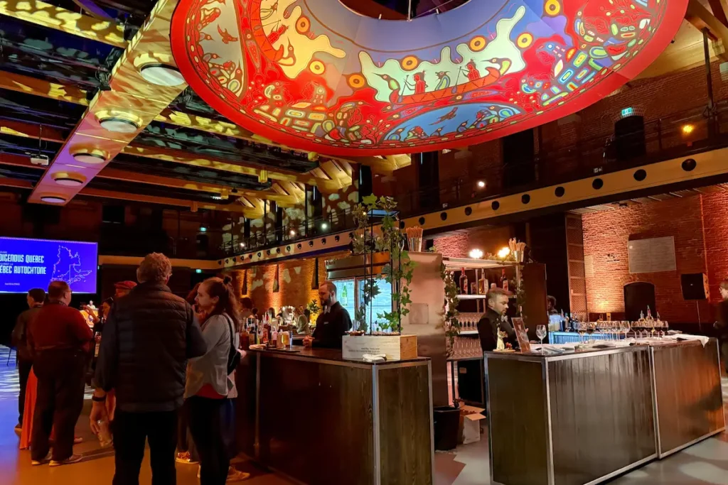 A warmly lit event space for TBEX closing reception party features a large circular ceiling mural in vivid red, blue, and gold with Indigenous art motifs, a bar area with drinks, and groups of people socializing.