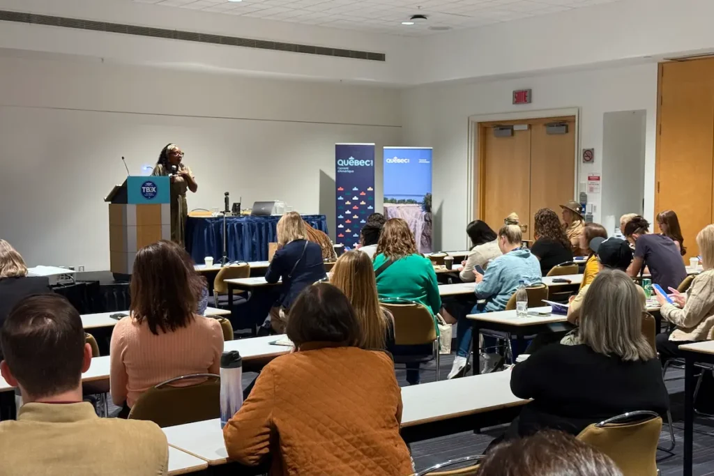 A TBEX session speaker presents at a podium in front of Québec tourism banners while attendees sit at tables, listening and taking notes.