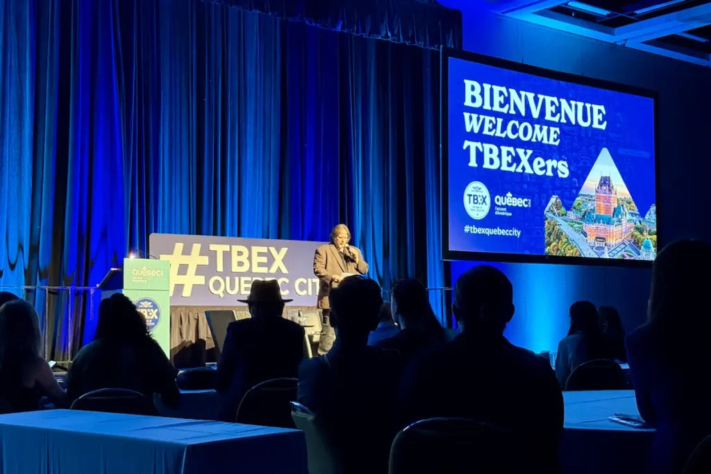 A man speaks on stage at TBEX Québec City under blue lighting, with a large screen reading “Bienvenue Welcome TBEXers” and an image of Château Frontenac.