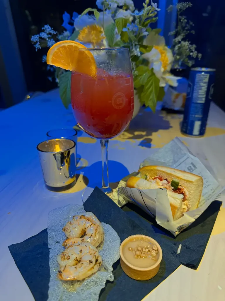 A table setting at the TBEX opening party displays grilled shrimp on a crisp wafer, a small dessert tart, a lobster roll, and a tall red cocktail garnished with an orange slice, with flowers in the background.