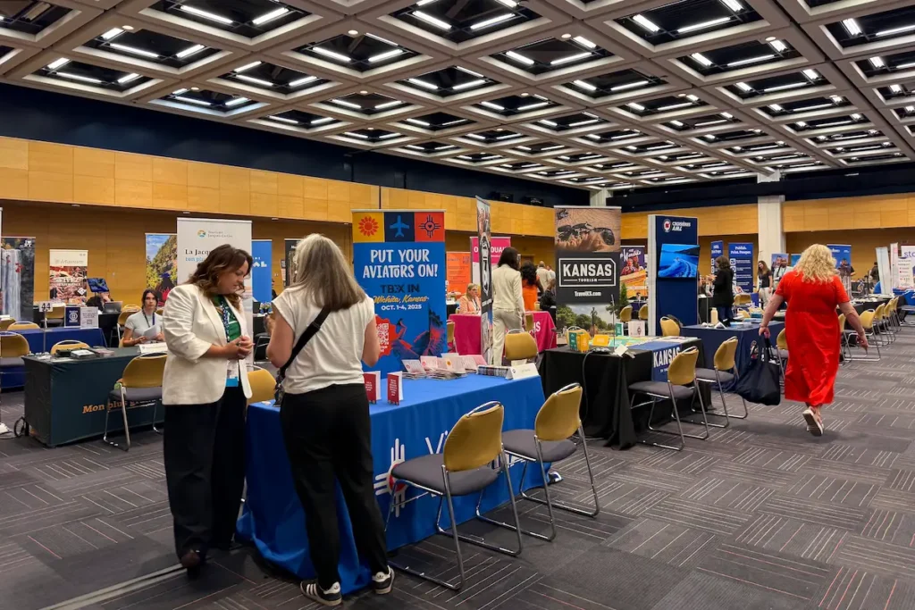 An exhibition hall during TBEX sessions features rows of tables showcasing tourism booths for various destinations, as attendees network and browse promotional displays.